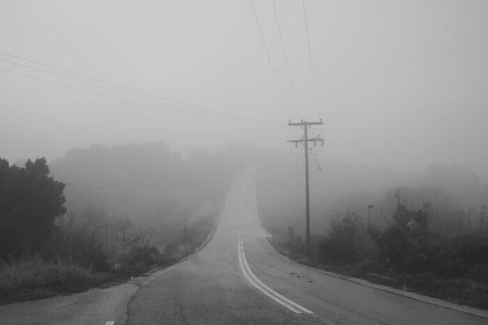 A winding road disappearing into thick fog, surrounded by colorful autumn foliage.