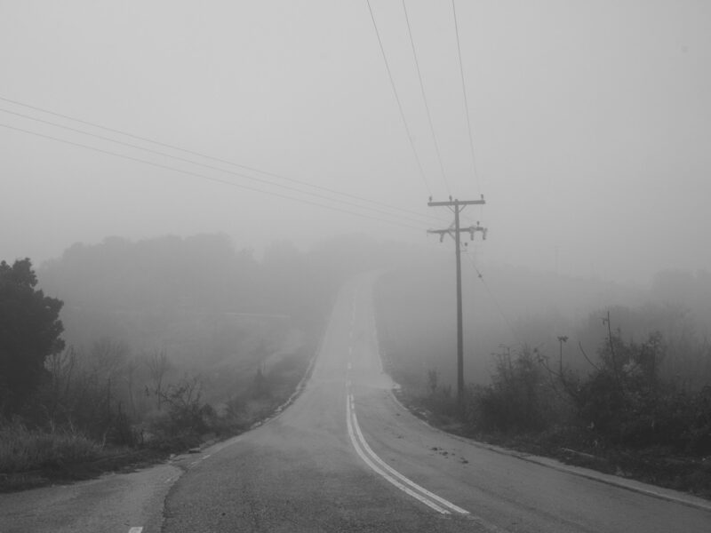 A winding road disappearing into thick fog, surrounded by colorful autumn foliage.