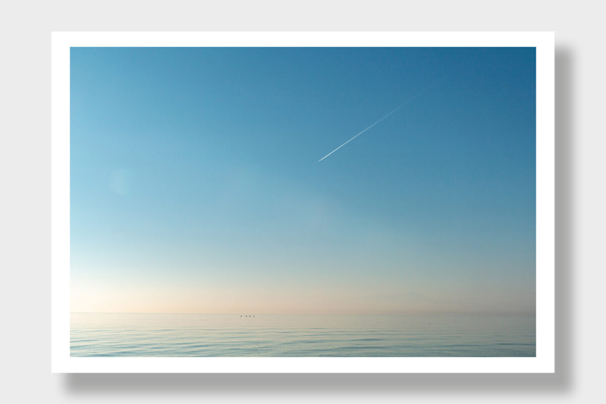 A serene scene in Greece: waveless turquoise sea, cloudless sky, and a streak resembling an airplane.