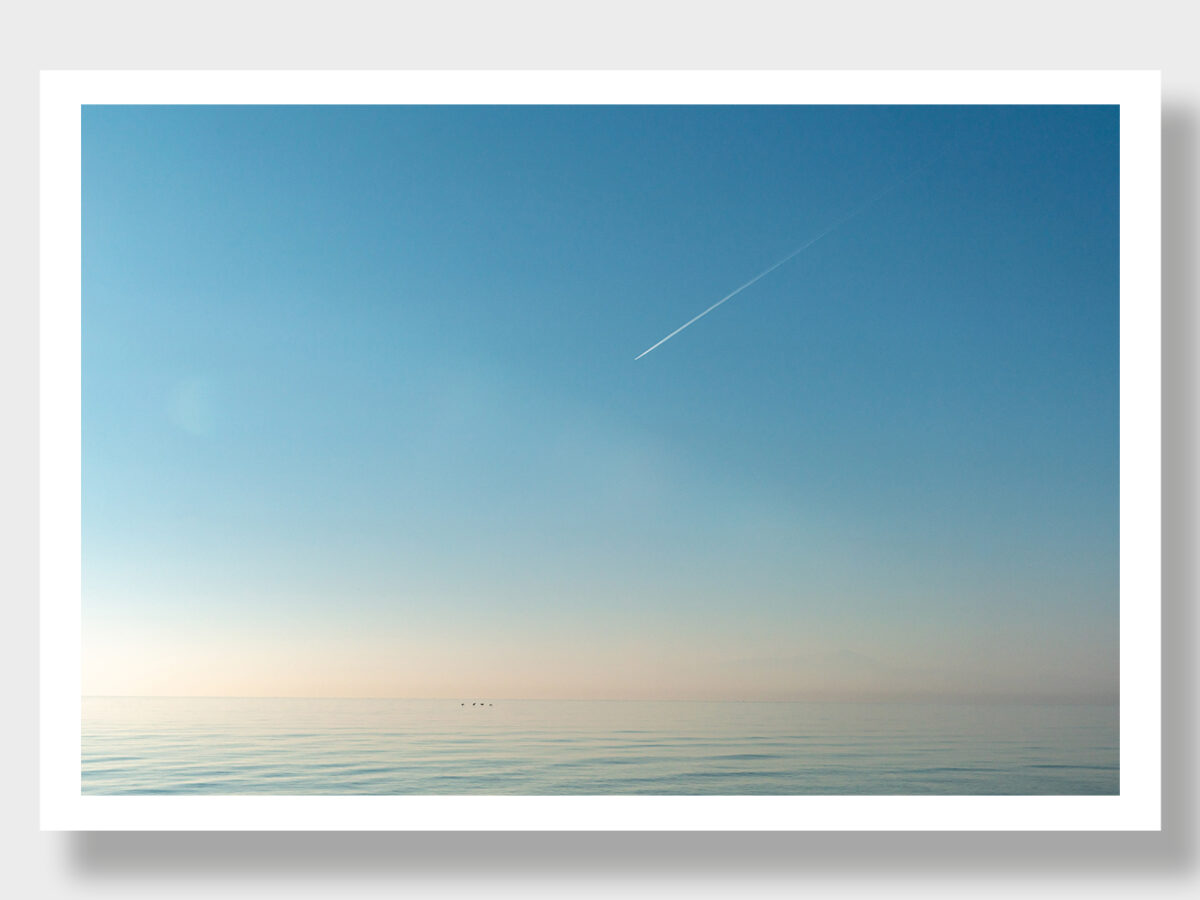 A serene scene in Greece: waveless turquoise sea, cloudless sky, and a streak resembling an airplane.